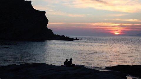 Young loving couple sitting by the sea in a beautiful bay with island view at sunset