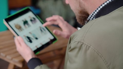 Shot from behing of a stylish guy looking at the tablet with open ecommerce website, man shopping online for summer clothes. Man choosing new clothes on internet. Blurred screen.