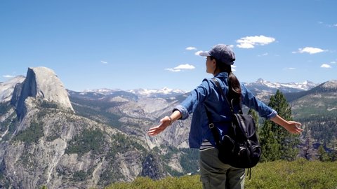 back view of young asian woman tourist arms raised enjoying fresh air top of mountain in half dome trail yosemite national park. happy female backpacker open hands standing on cliff rock blue sky