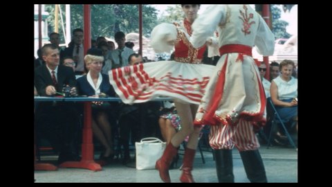 1960s: Men and women sit at judge's table. Couples in traditional Polish costumes dance at fairgrounds pavilion. Audience sits at tables and watches dancers.