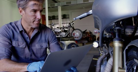 Front view close up of Caucasian male mechanic using laptop in motorbike repair garage. He is looking at motorbike 4k