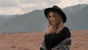 Close-up portrait: a beautiful young woman in a black hat stands in the open nature against the mountains, looking to the side and her hair develops wind. Slow motion, 4K