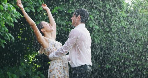 Slow motion of young carefree couple in love are dancing and kissing under the rain on a background of green trees.