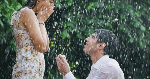 Slow motion of young handsome man is making a proposal of marriage to his fiancee under the rain on a background of green trees.
