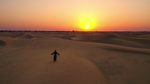 Aerial view from a drone flying next to a woman in abaya United Arab Emirates traditional dress rising her arms on the dunes in the desert of the Empty Quarter. Abu Dhabi, UAE.