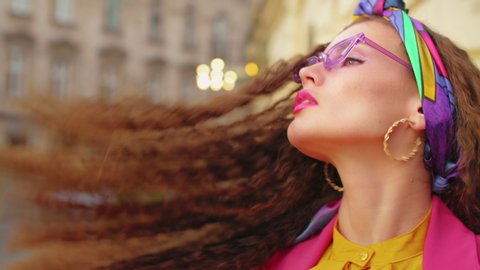 Close up portrait of fashionable woman with long curly hair, wearing colorful headband, purple sungasses, hoop earrings, wrist watch, rings, bold colors clothes, posing in street