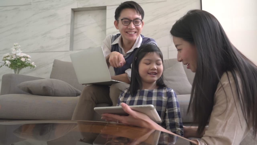slow motion asian daughter is doing a homework on the notebook. His mother and father are sitting and watch on sofa and use a digital laptop together home background.