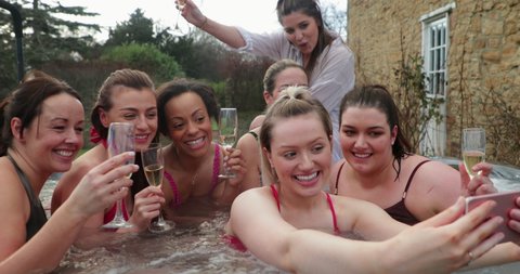 Small group of female adults celebrating with a glass of champagne while sitting in a hot tub. They are taking a selfie while relaxing together.