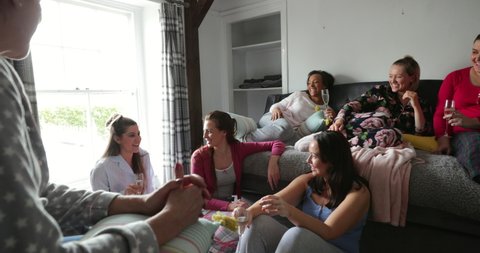 Over the shoulder view of a group of female friends sitting in their pyjamas in a bedroom. They are talking and having fun together.