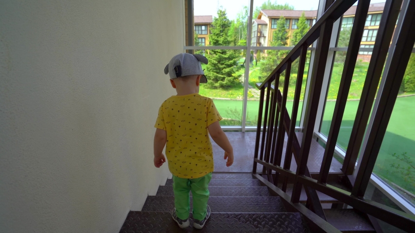 A little boy in a yellow t-shirt, light green pants and cap goes down the metal stairs, in a room with panoramic windows outside the window Sunny summer weather