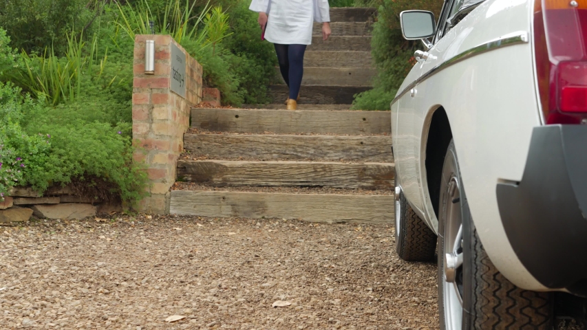Well dressed young lady walks towards camera, down stairs and past vintage car on farm