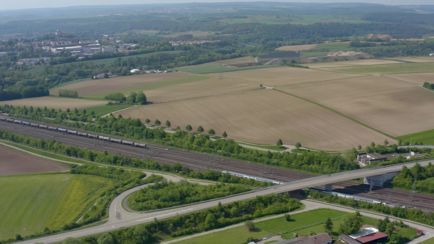 Aerial of a train driving by close to Ensingen.  Camera rotates right tracking train along railroad tracks, then tilts up. In foreground and background on either side of tracks are fields, wooded area