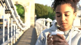 4K video clip of beautiful mixed race African American girl teenager young woman on a bridge over a river, drinking takeout coffee smiling, laughing and looking to camera