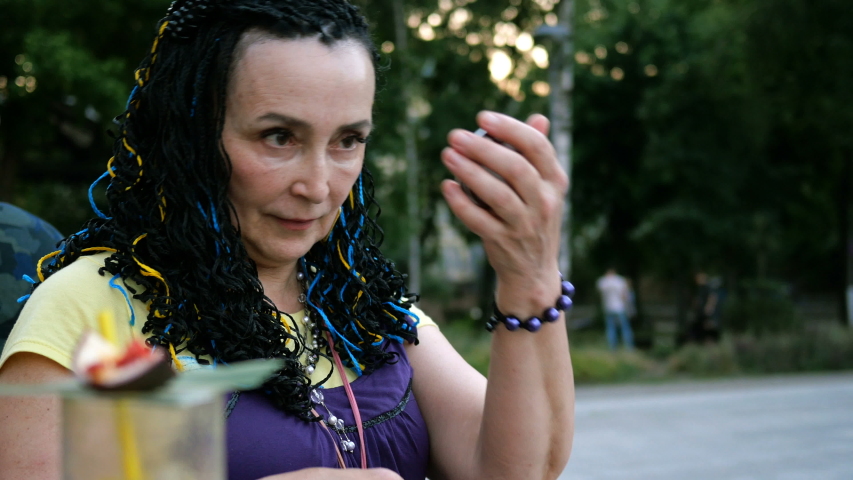 Senior extravagant woman sitting and straightens hair looking in a small mirror in the park among the trees. Outdoors. Close-up.