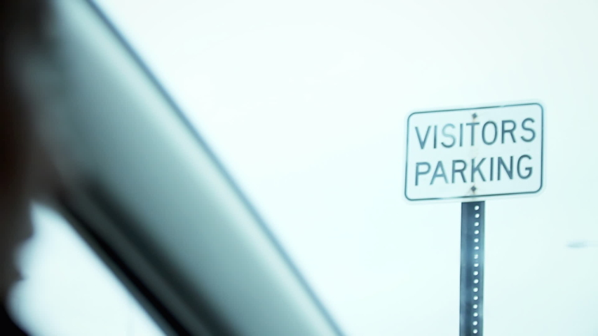Inside a car on a rainy day, looking at a sign with the words Visitors Parking. Handheld shot.