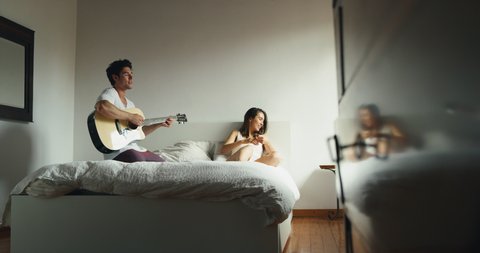 Authentic of young just married couple wearing pajamas having fun to playing guitar and singing in the bedroom in the early morning in a sunny day.