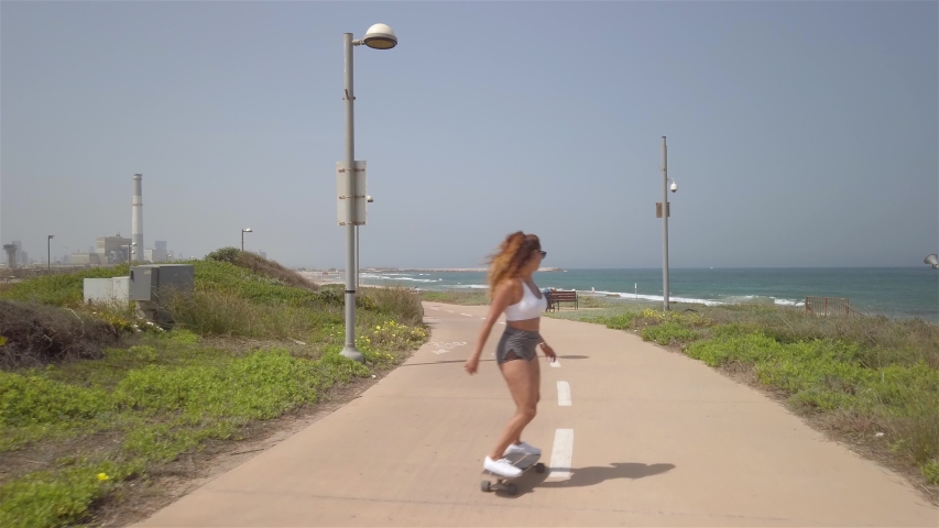 Young athletic woman surf-skating down the Tel aviv beach promenade on a bright sunny morning. Reading power station in the background. Following shot. 