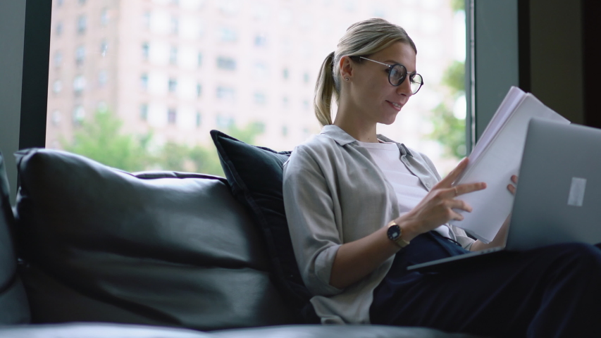 Young woman in eyeglasses reviewing paper documents and checking information during remote work at laptop in modern open space office.Hipster student doing homework and preparing coursework Royalty-Free Stock Footage #1031298491