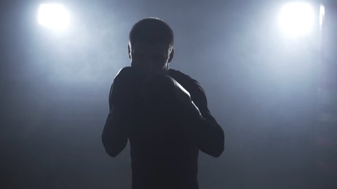 Kickboxer training in low light gym. Sportsman boxing in camera. Muay thai fighter punching. Silhouette on dark background. Medium shot with camera shaking when boxer punching