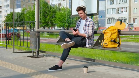 Joyful young man is waiting for transport at bus stop. male sitting on bench . Guy using smartphone listens music