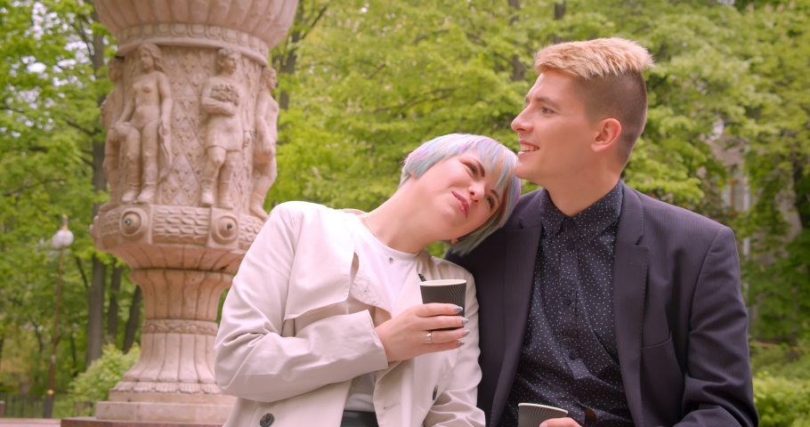 Closeup shoot of young beautiful hipster couple sitting and drinking coffee being happy in the park outdoors outdoors. Woman leaning on her husbands shoulder