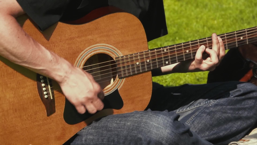 Close up of a young man playing acoustic guitar while sitting outside on the grass. Man wearing jeans and black shirt strumming a guitar in the sunshine.