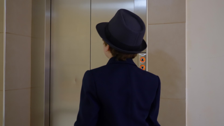 Little boy in business suit, hat and briefcase comes into elevator back view. Businessman pushing button for calling lift and entering into elevator car