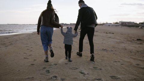 A young family walks along the beach near the sea on a windy day, the husband and wife carry a young son with his hands and make him fly. The couple enjoys a day off. Rare view
