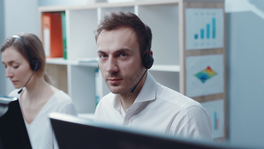 Face young handsome man operator look at camera smie taking calls smile in call center office business worker computer corporate headset job service group finance sunshine phone close up slow motion Royalty-Free Stock Footage #1031328632