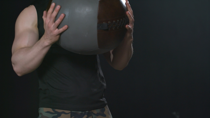 Close up shot of muscular young man holding medicine ball and performing weighted sumo squats in dark room
