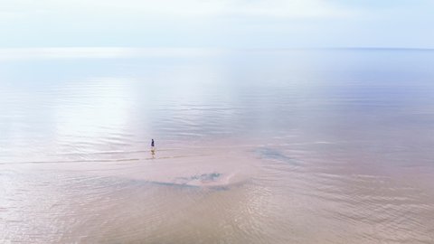 Aerial: Young woman in blue bright dress walking around - Shallow water scenic yellowish sunset landscape at the Baltic Sea - Drone flying view from above