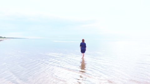 Aerial: Young woman in blue bright dress walking around - Shallow water scenic yellowish sunset landscape at the Baltic Sea - Drone flying view from above
