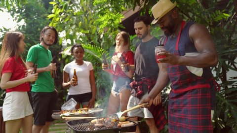 Group of people standing around barbecue grill, chatting, drinking and eating at summer outdoor party and holidays concept. Adlı Stok Video