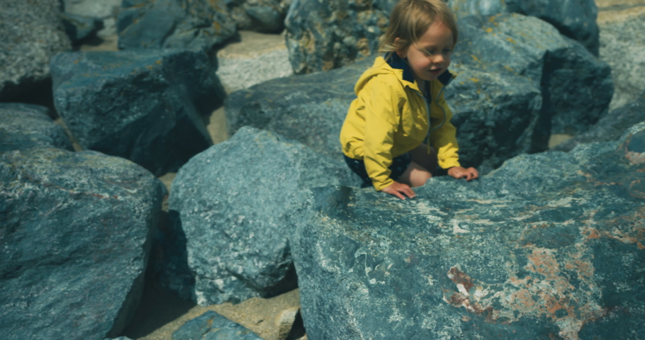 A little toddler is climbing the rocks on a beach with his grandfather relaxing nearby
