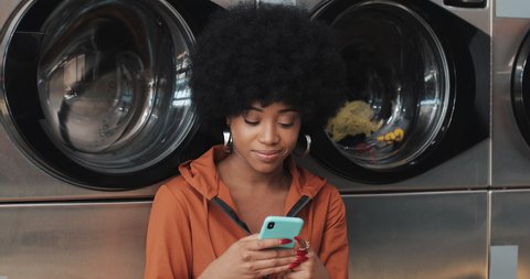 Smiling young woman using smartphone at laundromat. Self-service public laundry.