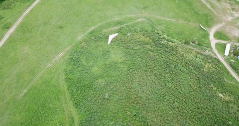 Flying on a light hang glider. Shooting from a drone, top view. Paraglider flying over green meadows. See the path and the bushes.
