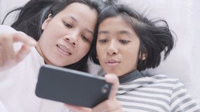 Mom and daughter are watching funny clips together. Asian women and girl are cute, fun and happy on the bed at home.