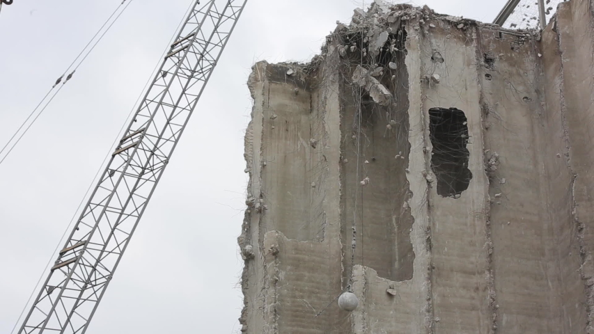 Wrecking ball hanging from crane chain near building under demolition. Long shot.