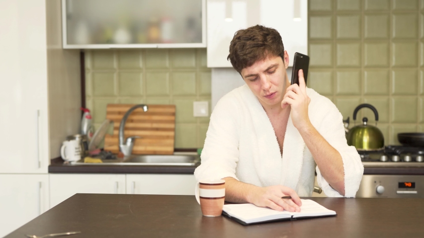 Young handsome man is sitting in a kitchen and talking on the phone. His beautiful wife give him a cup of tea and a croissant.