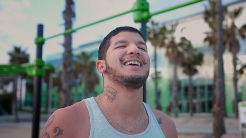 Portrait of attractive Hispanic guy smiling and standing after hard training at beach, close up of bearded funny sportive male enjoying rest after his morning workout at modern urban spot in Spain