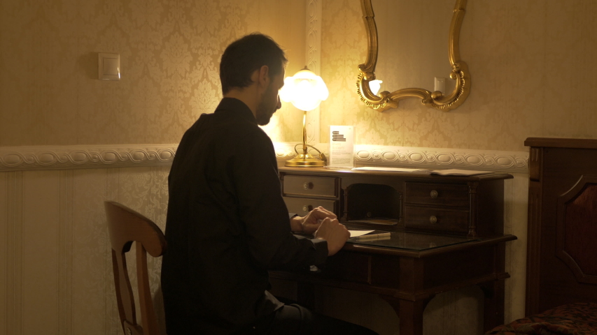 Man in black shirt takes out of a drawer his glasses, clean them and start writing in a dark room with a table light.