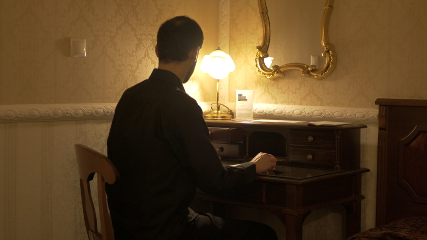 Man wearing black shirt in a dark room with a table light is wearing his glasses and writes a letter on a wooden desk in front of a mirror.