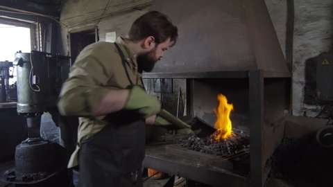 Back view of Caucasian bearded blacksmith in apron and gloves approaching to stove with shovel in his hands, taking new coals and putting them into stove then moving damper to blow fire up