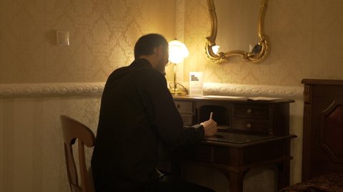 Man in black shirt is writing something in a dark room with a table light.