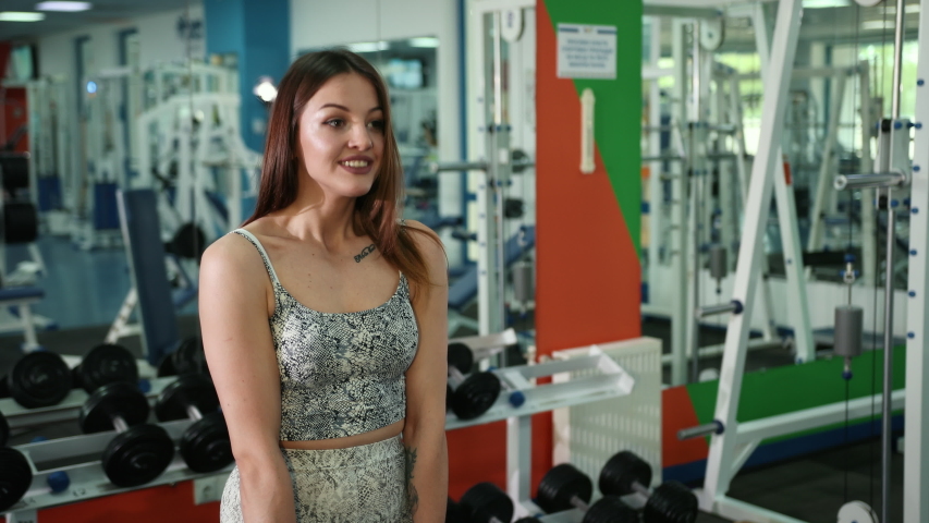 A cute, chic young woman raises dumbbells bending her body in the gym. Dressed in a gray suit. Behind the mirror. Long dark hair and brown eyes. Model appearance. Prores, Slow Motion, 4k