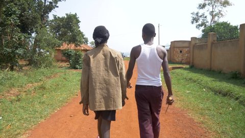 A tracking shot from behind of two young African gangsters with weapons walking down a dirt road in rural Africa.