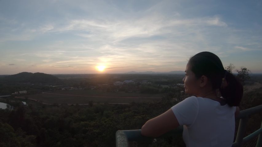 A slow tracking shot from left to right focusing on the back of a young asian girl standing at a high viewpoint admiring the landscape during the sunset