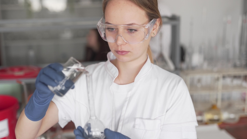Young Caucasian Female Scientist in Laboratory Wearing White Uniform Protective Glasses and Gloves Moving Liquid from Glass to Volumetric Flask. Blurred Background and Light Camera Movement