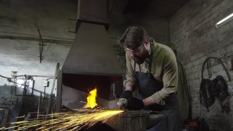 Tracking right of bearded Caucasian blacksmith in safety apron, gloves and glasses standing at anvil and shaping iron detail using hand electrical circular saw with abrasive disk
