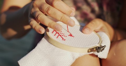 Unrecognizable woman embroidering beautiful red patterns. 4k close up macro shot of female hands sewing cross-stitch outside on nature background slow motion. Needlework handicraft business lifestyle
